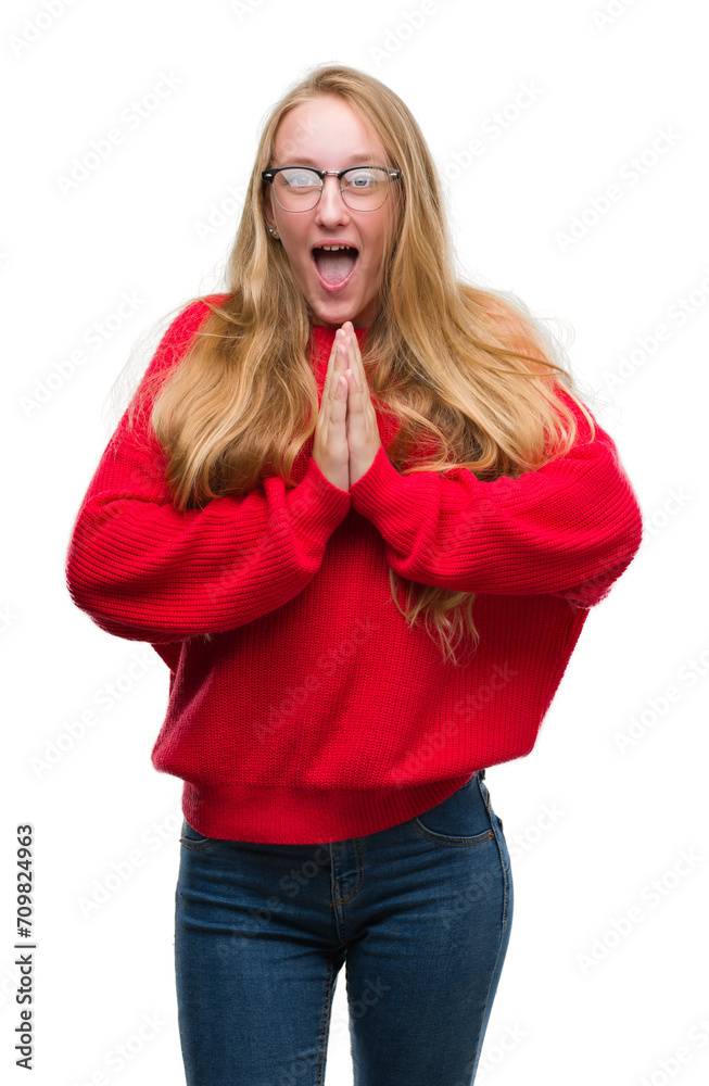 Blonde teenager woman wearing red sweater begging and praying with hands together with hope expression on face very emotional and worried. Asking for forgiveness. Religion concept. Blonde teenager woman wearing red sweater begging and praying with hands together with hope expression on face very emotional and worried. Asking for forgiveness. Religion concept.