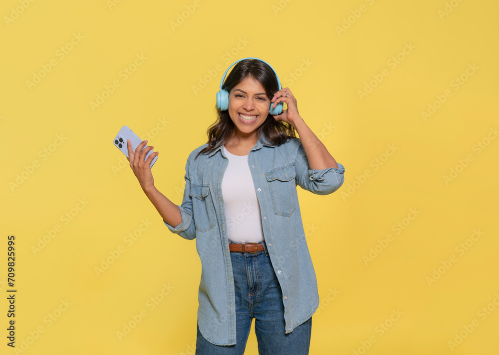 Happy Indian woman carried away with music dancing carefree wearing headphones on her ears dressed in jeans and denim shirt, looking at camera, using phone isolated on a yellow background Happy Indian woman carried away with music dancing carefree wearing headphones on her ears dressed in jeans and denim shirt, looking at camera, using phone isolated on a yellow background