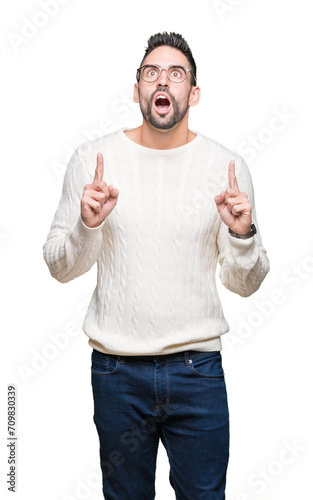Young handsome man wearing glasses over isolated background amazed and surprised looking up and pointing with fingers and raised arms.