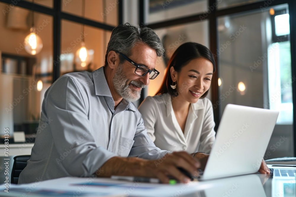 Mature Latin manager mentor talking to young Asian female coworker, showing online project results at meeting. Two happy diverse professional executives team working in office using pc, Generative AI