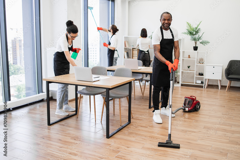 Young adult African American man wearing black apron and red rubber gloves vacuuming floor of modern light office. Professional service team of cleaning company. Young adult African American man wearing black apron and red rubber gloves vacuuming floor of modern light office. Professional service team of cleaning company.