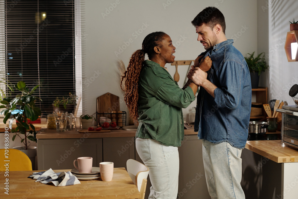 Side view of young furious woman shouting at her husband and blaming him while holding collar of his shirt during argument Side view of young furious woman shouting at her husband and blaming him while holding collar of his shirt during argument