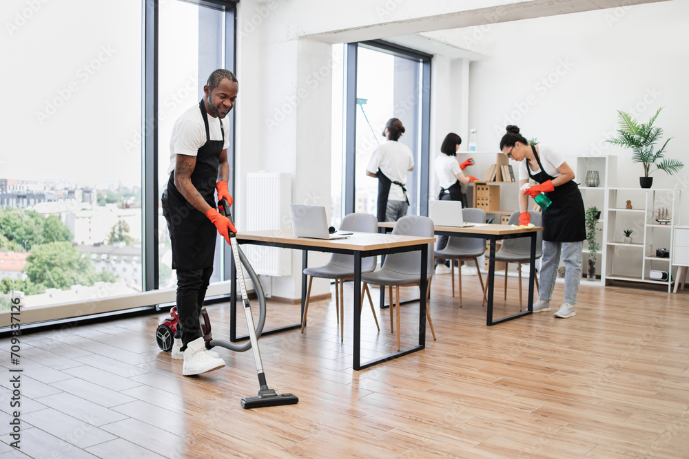 Professional service team of cleaning company. Young adult African American man wearing black apron and red rubber gloves vacuuming floor of modern light office.