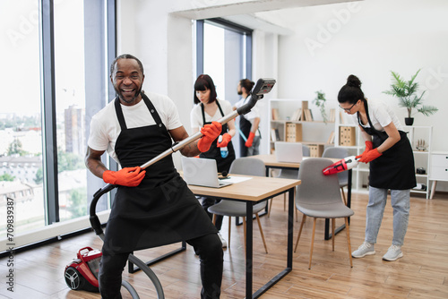 Happy multinational team of people in black aprons cleaning modern office. Professional cleaner young African man having fun holding vacuum cleaner like guitar imitating playing at concert.