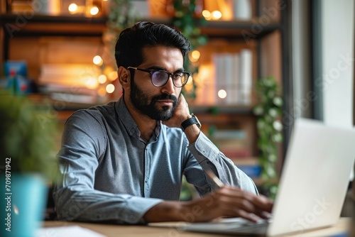 Young indian professional business man, focused ethnic male student wearing glasses working on laptop, remote studying using computer looking at screen watching seminar webinar at home, Generative AI