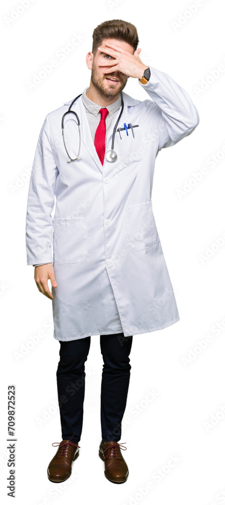 Young handsome doctor man wearing medical coat peeking in shock covering face and eyes with hand, looking through fingers with embarrassed expression. Young handsome doctor man wearing medical coat peeking in shock covering face and eyes with hand, looking through fingers with embarrassed expression.