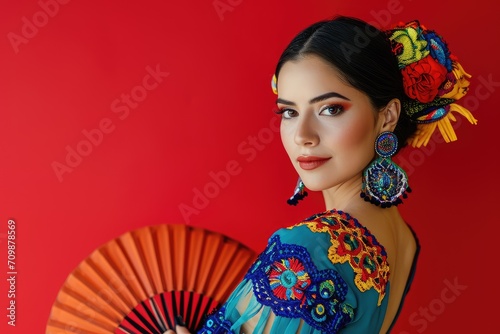 Studio portrait of a Latina woman in a colorful flamenco dress, with a traditional fan, against a vibrant red background.