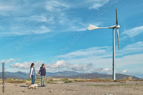 Green energy concept. Couple walking with dog near mountain top industrial wind power plant, wind turbine for generating electricity in wind farm or wind park.