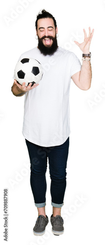 Young man with long hair and bear holding soccer football ball very happy and excited  winner expression celebrating victory screaming with big smile and raised hands