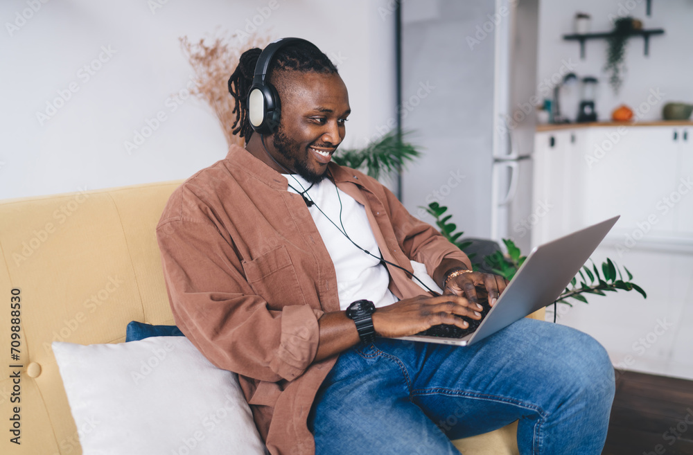 Cheerful black man in headphones typing while using laptop in living room