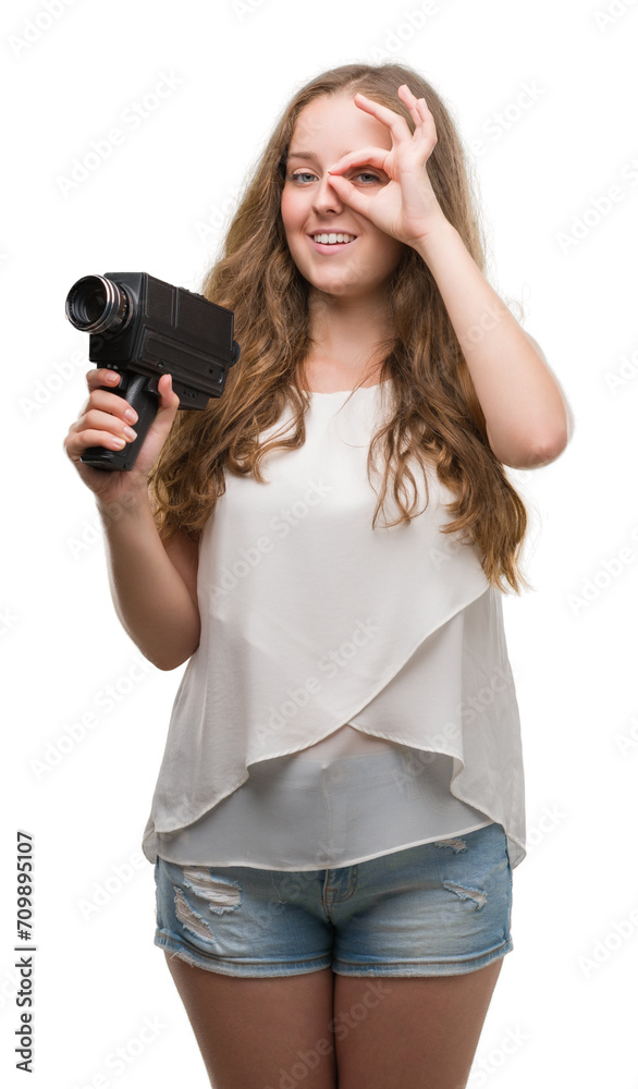 Young blonde woman holding super 8 video camera with happy face smiling doing ok sign with hand on eye looking through fingers Young blonde woman holding super 8 video camera with happy face smiling doing ok sign with hand on eye looking through fingers