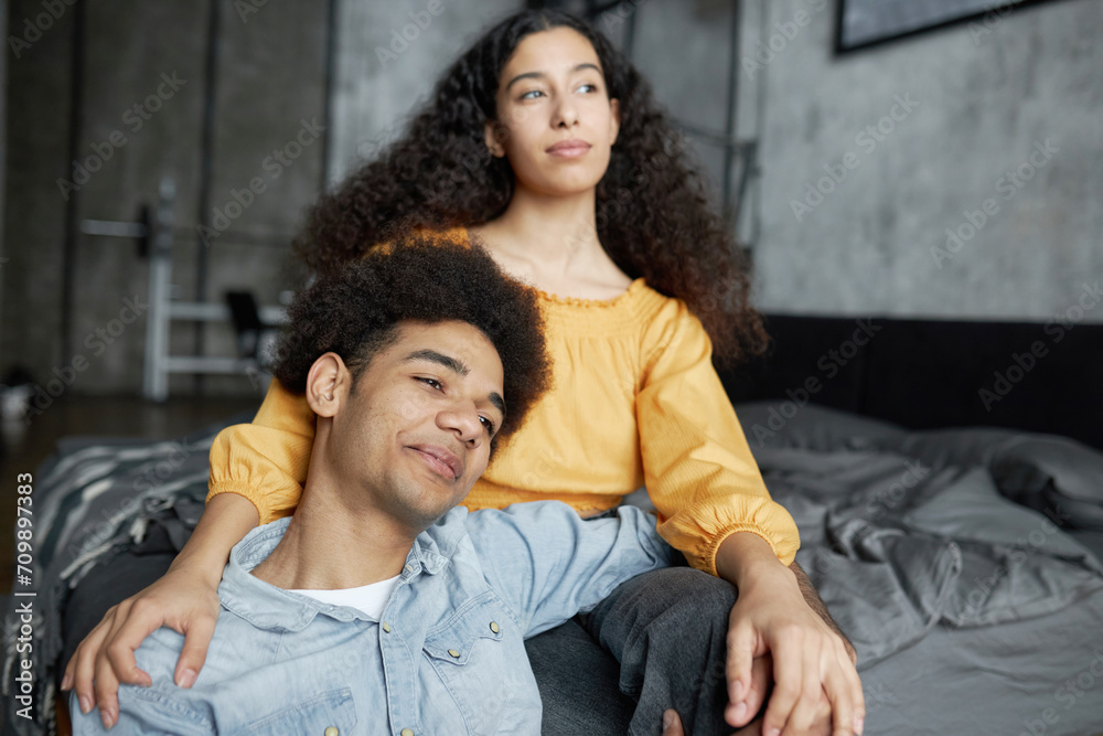 Young black couple spending time together, man sitting on floor next to his girlfriend on bed, hugging her legs, smiling with dreamy face, female looking aside with sly thoughtful facial expression