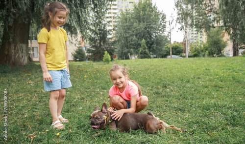 Bulldog lies in the grass. Two preschool girls play with a bulldog dog. Children and animals on a walk. Communication between children and animals.
