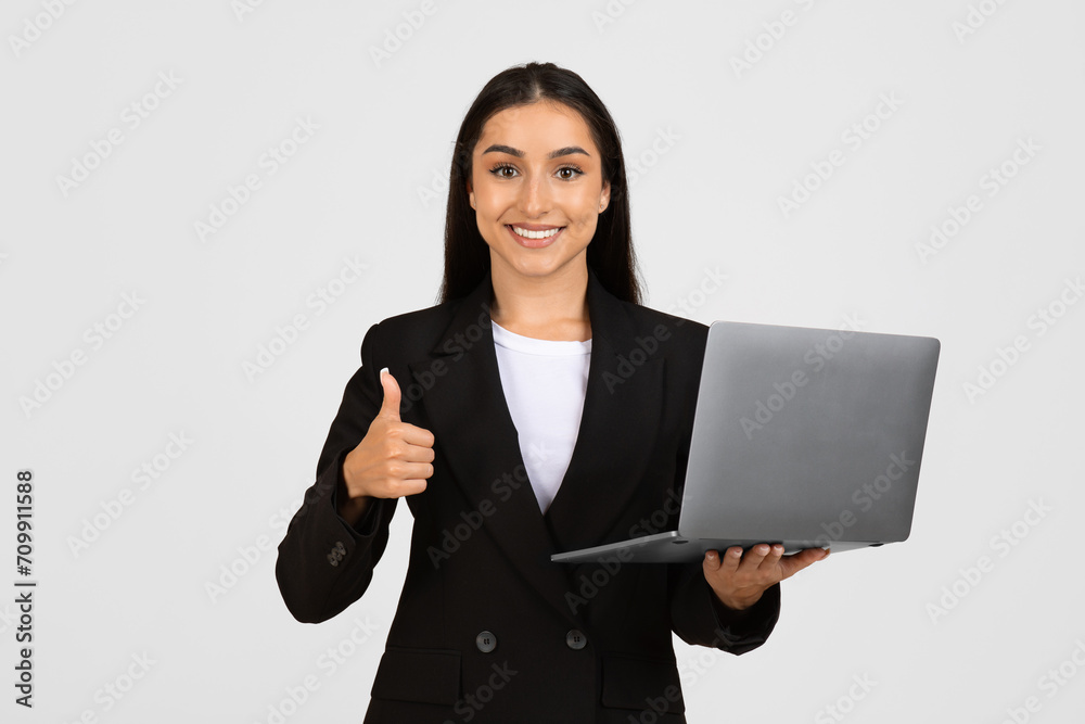 Glad european business lady in suit, with computer, making thumb up gesture, isolated on grey studio background Glad european business lady in suit, with computer, making thumb up gesture, isolated on grey studio background