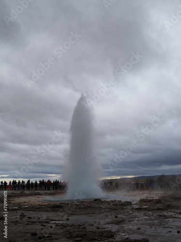 Haukadalur geothermal valley - geysir eruption with water and steam in the middle, surrounded by tourists, under a cloudy sky