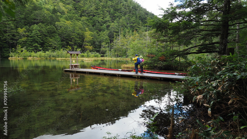 A pair of travelers in colorful hiking clothes visiting traditional Japanese shrine at Myojin Pond, Kamikochi, Japan, with mirror image of surrounding forest, mountains and sky in the lake