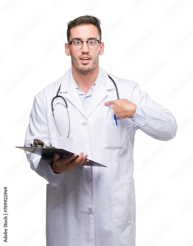 Handsome young doctor man holding a clipboard with surprise face pointing finger to himself