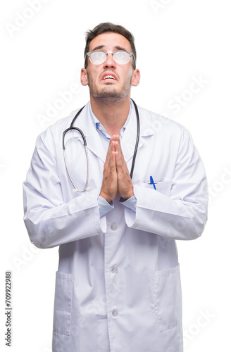 Handsome young doctor man begging and praying with hands together with hope expression on face very emotional and worried. Asking for forgiveness. Religion concept.