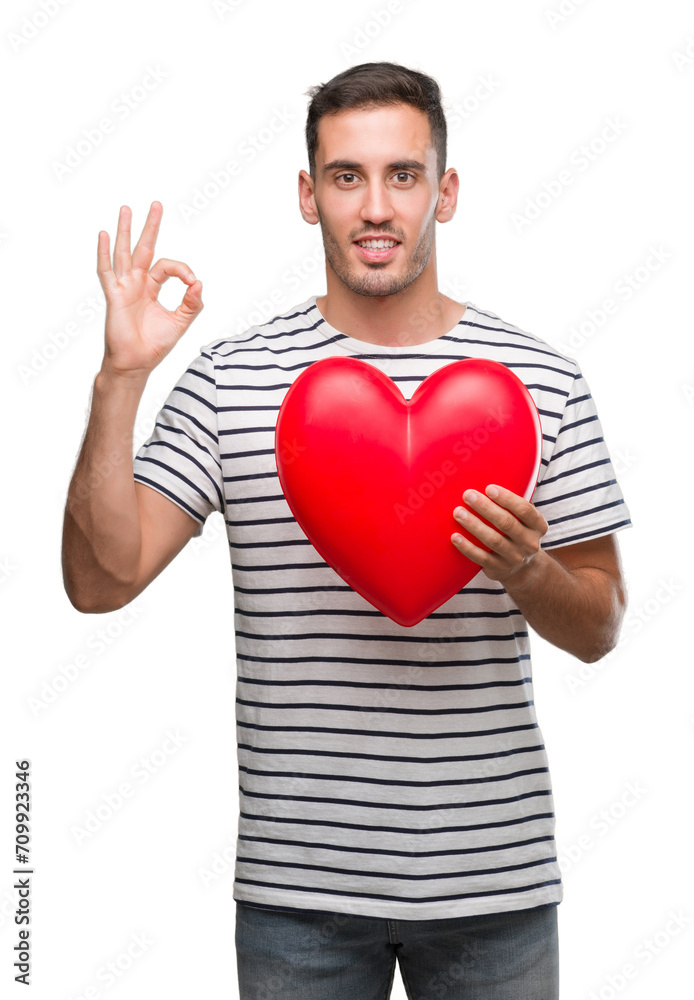 Handsome young man in love holding red heart doing ok sign with fingers, excellent symbol Handsome young man in love holding red heart doing ok sign with fingers, excellent symbol