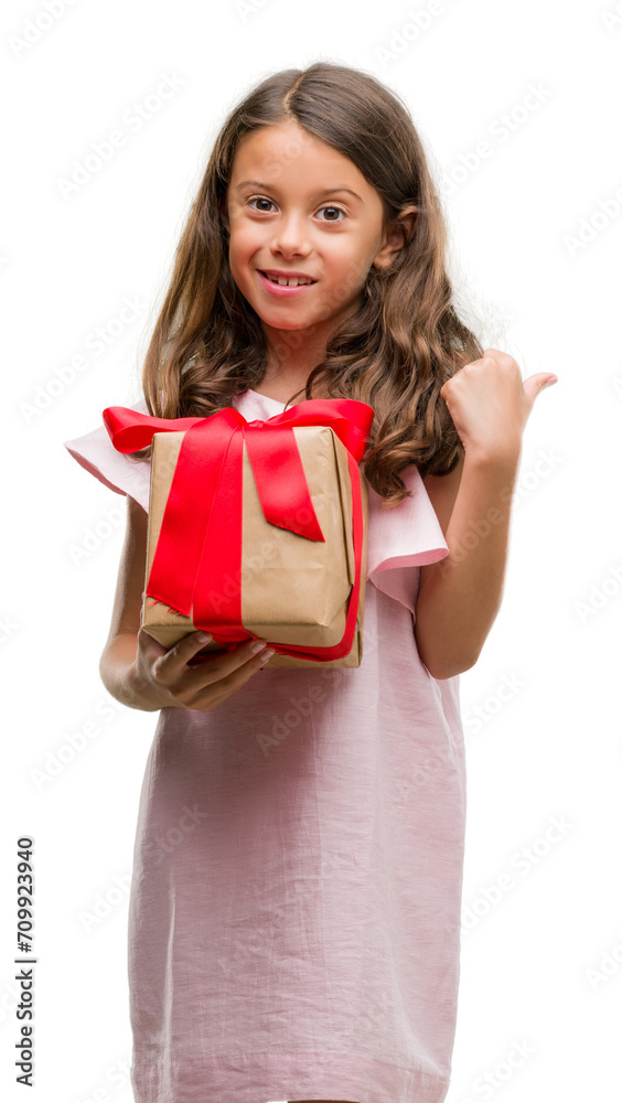 Brunette hispanic girl holding a gift pointing and showing with thumb up to the side with happy face smiling Brunette hispanic girl holding a gift pointing and showing with thumb up to the side with happy face smiling
