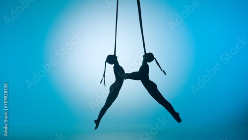 Silhouette of two female acrobats isolated on blue neon background. Girls aerial dancers performing acrobatic flying on ropes.