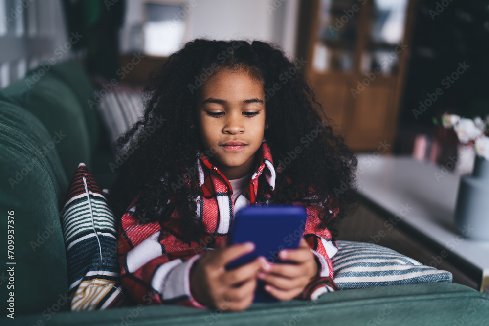 Fototapeta premium Young African-American girl engaged with her handheld smartphone device, comfortably snuggled on a couch at home, showing a moment of modern childhood leisure