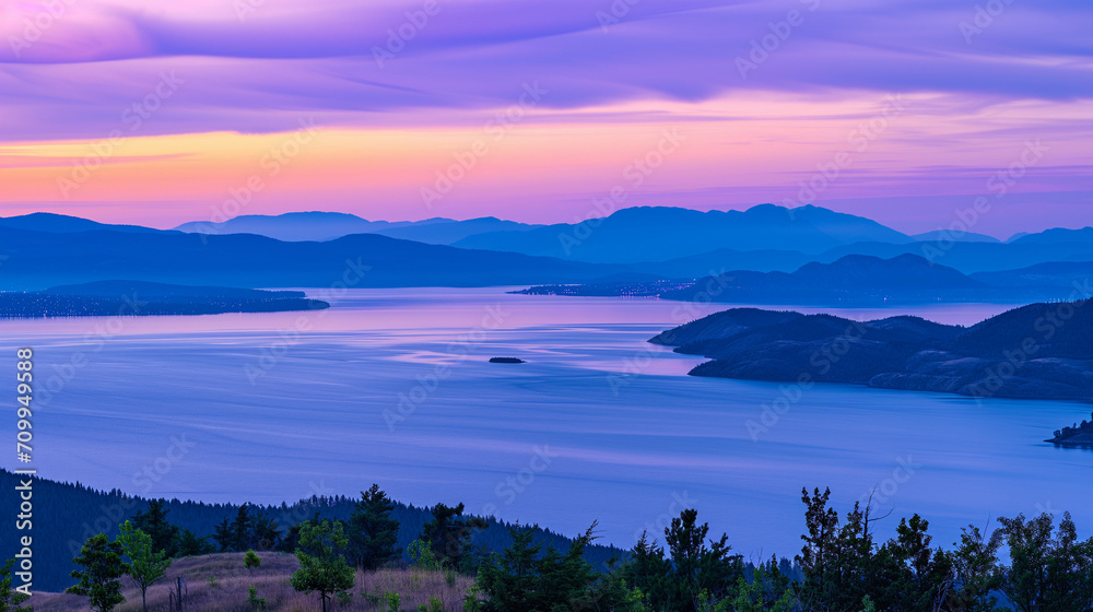 An aerial view of a coastline with distant lights from a small settlement, set against mountain ranges during dusk, casting vibrant shades of purple and blue across the landscape