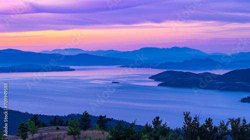An aerial view of a coastline with distant lights from a small settlement, set against mountain ranges during dusk, casting vibrant shades of purple and blue across the landscape