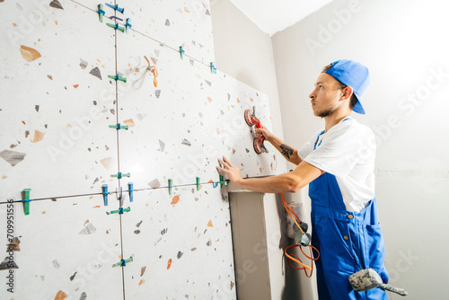 Adult repairman in a special uniform laying tiles on the wall at bathroom in new house 