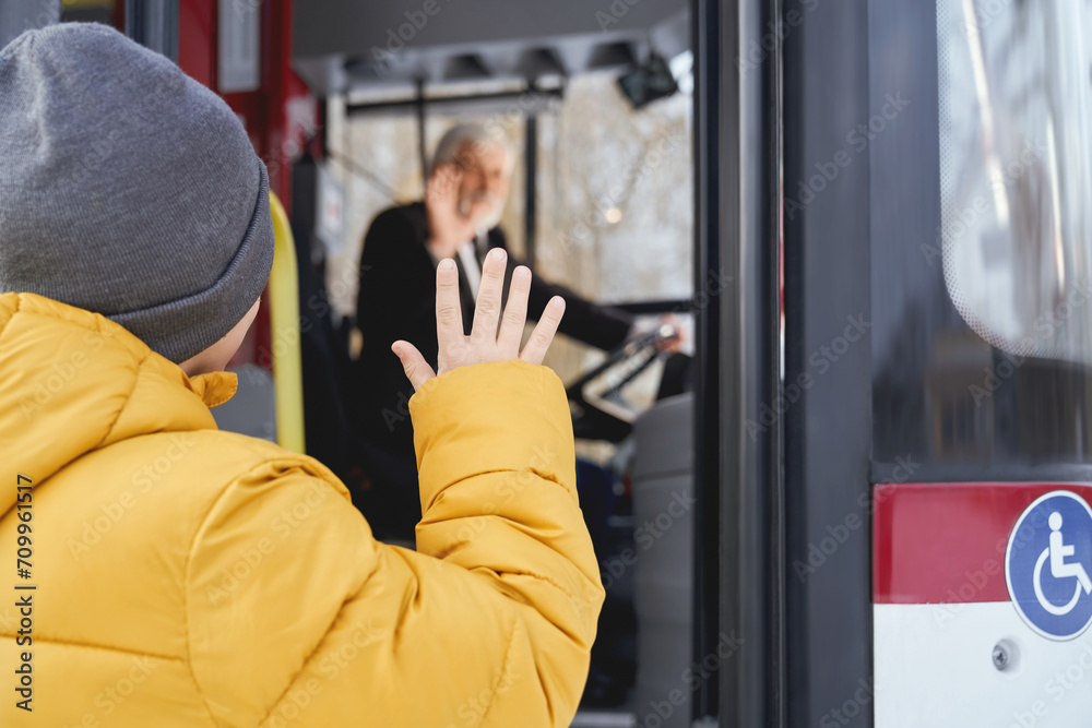 Back view of little boy in orange jacket and grey cap saying goodbye to blur driver after travelling in public vehicle. Crop of child male waving to driver while getting off bus. Concept of city life. Back view of little boy in orange jacket and grey cap saying goodbye to blur driver after travelling in public vehicle. Crop of child male waving to driver while getting off bus. Concept of city life.