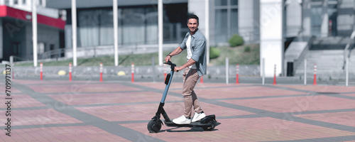 Summer adventure. An effortlessly stylish, slim young adult man joyfully rides an electric scooter with a beaming smile across a sunlit city square, surrounded by contemporary buildings