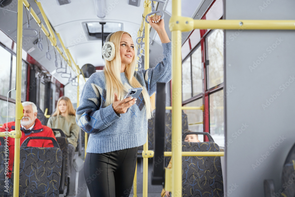 Side view of stylish blonde girl with phone looking at window smiling standing inside moving public transport. Crop of female holding handle with blur passengers sitting in bus. Concept of city life. Side view of stylish blonde girl with phone looking at window smiling standing inside moving public transport. Crop of female holding handle with blur passengers sitting in bus. Concept of city life.