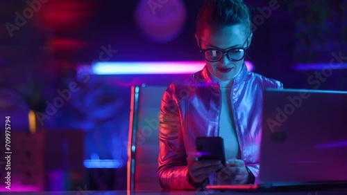 Neon metaverse futuristic concept. elegant woman in glasses with laptop using smartphone in modern office. photo