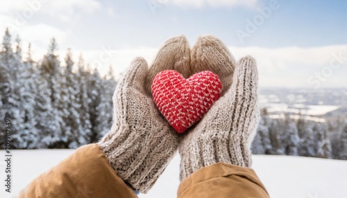 hands in gloves holding heart shape knitted object on a blurred winter landscape background with trees