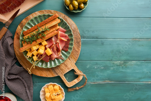 Slices of prosciutto or jamon. Antipasto Delicious grissini sticks with prosciutto, cheese, rosemary, olives on green plate on old wooden blue background. Appetizers table with italian snacks Top view photo