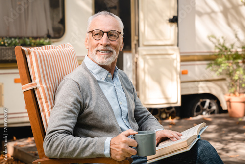 Closeup photo of old senior elderly grandfather man adventurer explorer relaxing in a chair reading book while traveling caravanning in motor home camper van trailer