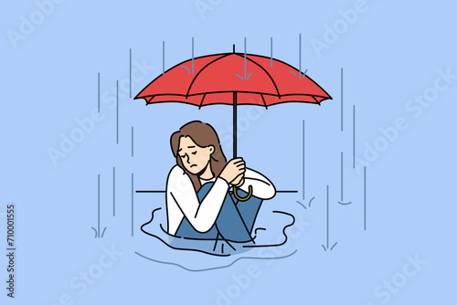 Woman sits in puddle in rain and holds umbrella over head, trying to escape autumn downpour. Homeless, wet girl is sad and apothetically looks away during rain, due to lack of home or place in shelter