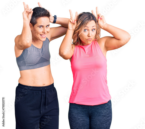 Couple of women wearing sportswear posing funny and crazy with fingers on head as bunny ears, smiling cheerful