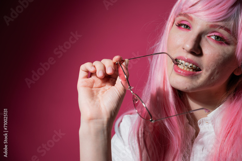 Close-up portrait of a young woman with braces in a pink wig and sunglasses on a pink background.