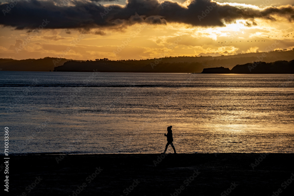Sunset beach scene, people silhouetted against a shimmering sea in the golden sunlight.
Families and beachgoers walk along in the evening-afternoon sunlight with a dramatic sky.
