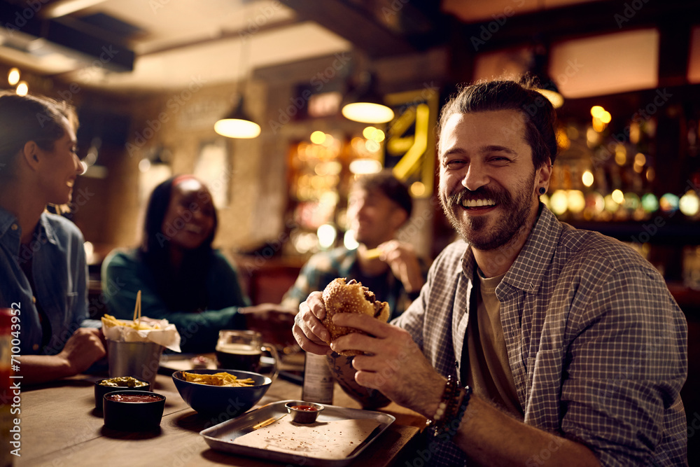 Happy man eating hamburger in pub and looking at camera.