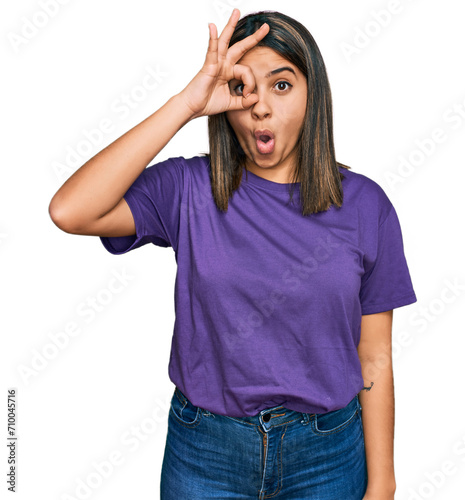 Young hispanic girl wearing casual purple t shirt doing ok gesture shocked with surprised face, eye looking through fingers. unbelieving expression.