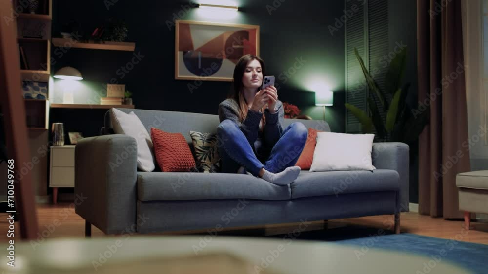 Woman relaxing sitting at sofa. Female using social media. Woman holding phone and smiling. Female using device for communication. Woman taking rest at home. Female touching smartphone screen.