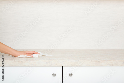 Young woman hand holding dry white paper napkin and wiping beige stone tabletop above kitchen cabinets at home. Closeup. Cleaning service. Front view. Empty place for text on wallpaper background.