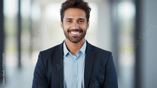 A professional young man with a charming smile stands confidently in a business environment, showcasing a smart casual look with a blue shirt and dark blazer