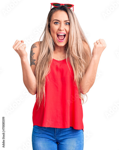 Young beautiful blonde woman wearing sleeveless t-shirt and sunglasses celebrating surprised and amazed for success with arms raised and open eyes. winner concept.