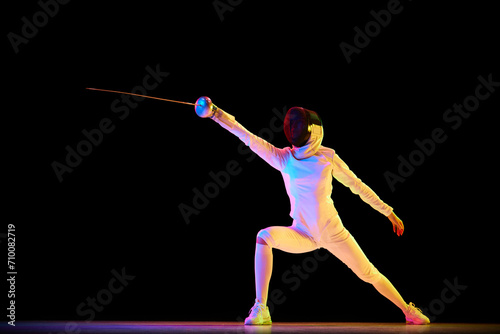 Female fencer, athlete in full uniform training, competing, practicing over black background in neon light. Concept of professional sport, competition, championship, hobby
