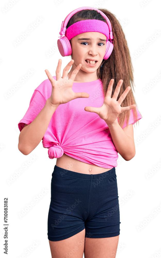 Cute hispanic child girl wearing gym clothes and using headphones disgusted expression, displeased and fearful doing disgust face because aversion reaction. with hands raised Cute hispanic child girl wearing gym clothes and using headphones disgusted expression, displeased and fearful doing disgust face because aversion reaction. with hands raised