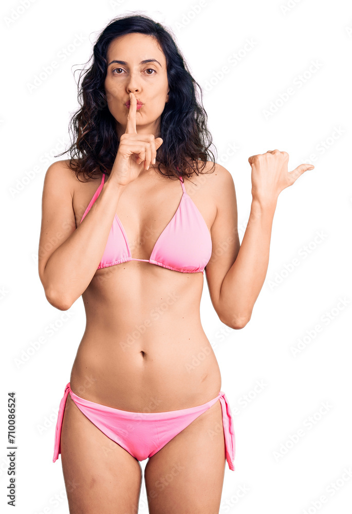 Young beautiful hispanic woman wearing bikini asking to be quiet with finger on lips pointing with hand to the side. silence and secret concept. Young beautiful hispanic woman wearing bikini asking to be quiet with finger on lips pointing with hand to the side. silence and secret concept.