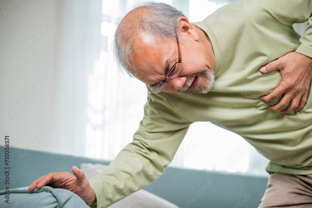 Obraz premium Sad senior man feeling bad pain use hand touching chest having heart attack on sofa in living room, Worried Asian old man have congenital disease suffering from heartache alone at home his heart aches